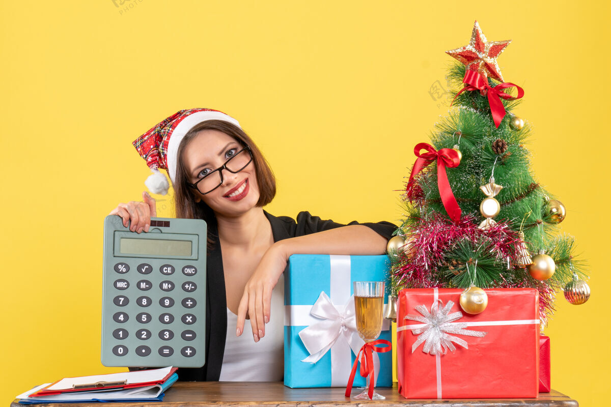 显示满意迷人的女士穿着西装 戴着圣诞老人的帽子 在办公室里展示计算器 黄色隔离帽子快乐圣诞老人