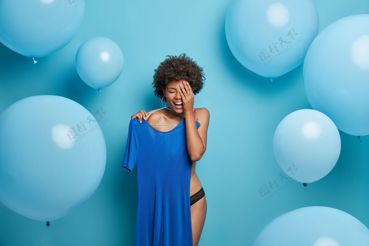 女人快乐的脱衣美国黑人年轻女子把礼服挂在衣架上 为蓝色主题的婚礼盛装 开怀大笑 对着蓝色的墙壁摆姿势 大气球单色镜头商场穿着服装