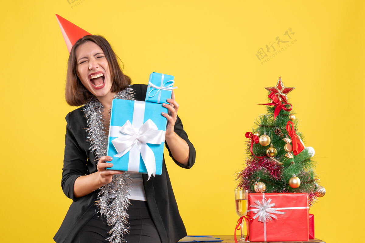 举行圣诞节心情动情美丽的女士戴着圣诞帽拿着礼物在黄色的办公室里花束礼物人