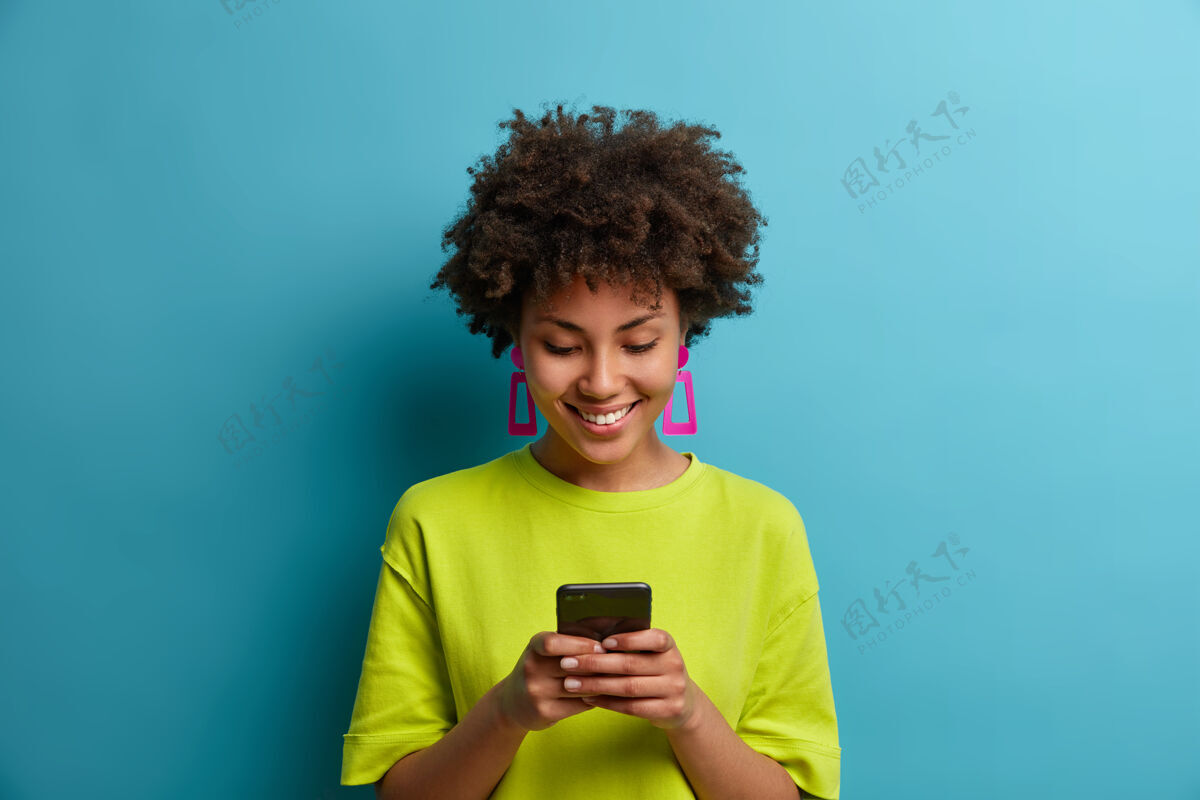 使用快乐快乐的卷发女人手持手机和社交网络中的朋友发短信 使用特殊的应用程序 观看有趣的视频 隔离在蓝色的墙上人和技术非洲非洲手机
