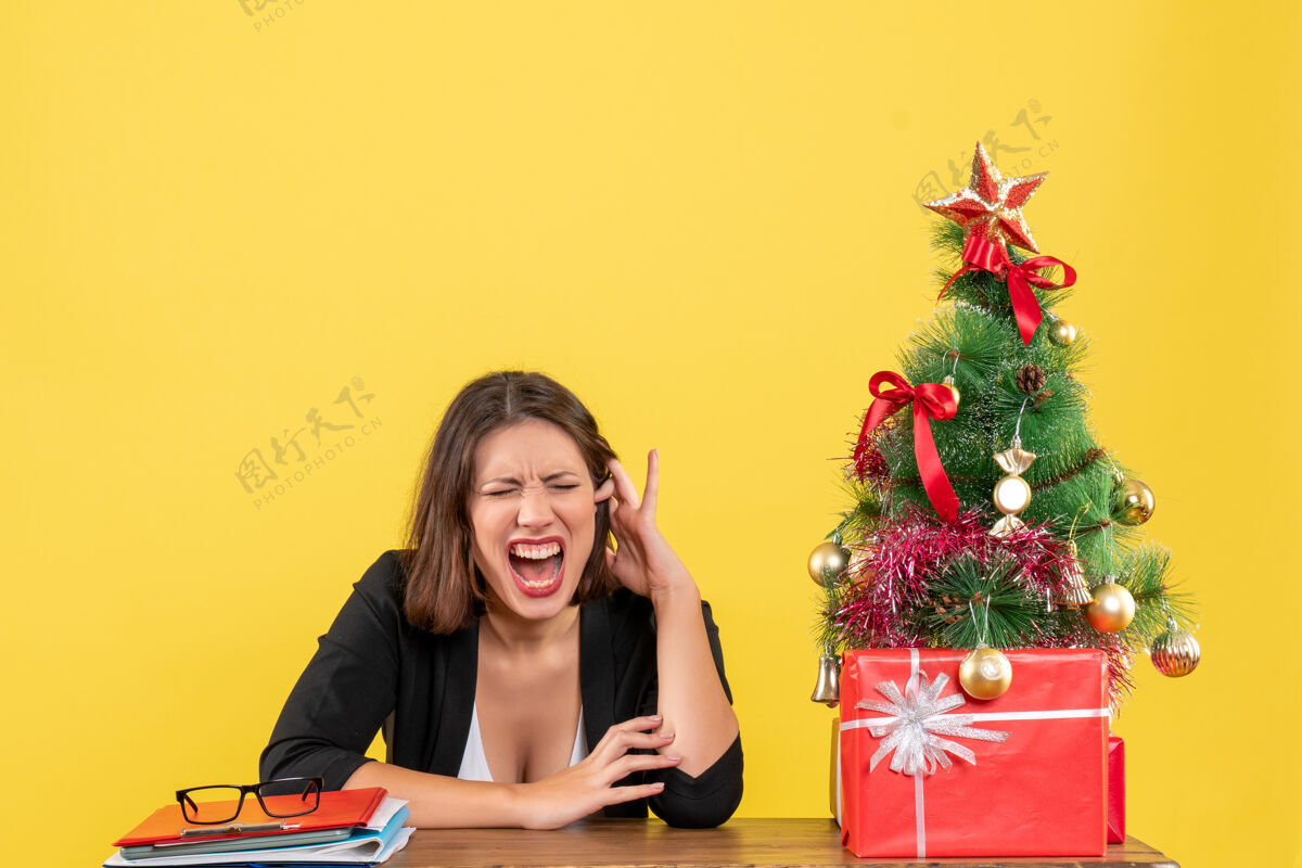 肖像情绪激动的年轻女子闭上眼睛坐在黄色办公室装饰圣诞树旁的桌子旁年轻女人坐着情绪