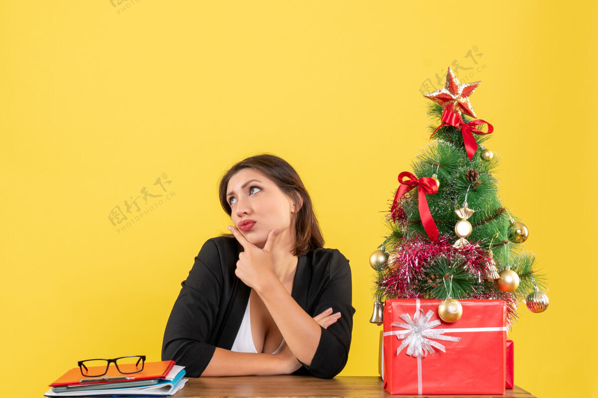 人年轻漂亮的女人正坐在黄色办公室装饰的圣诞树旁的桌子旁仔细思考着什么圣诞靠近年轻