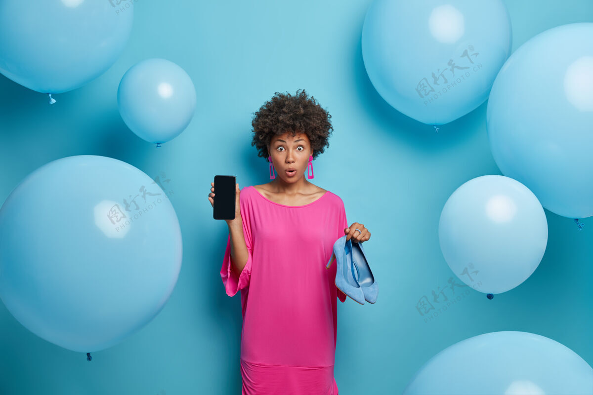 惊讶一张令人惊讶的卷发年轻女子的照片展示了手机显示屏和高跟鞋 网上购物 在网店买衣服 靠着蓝色的墙壁站着 周围有气球年轻鞋子衣服