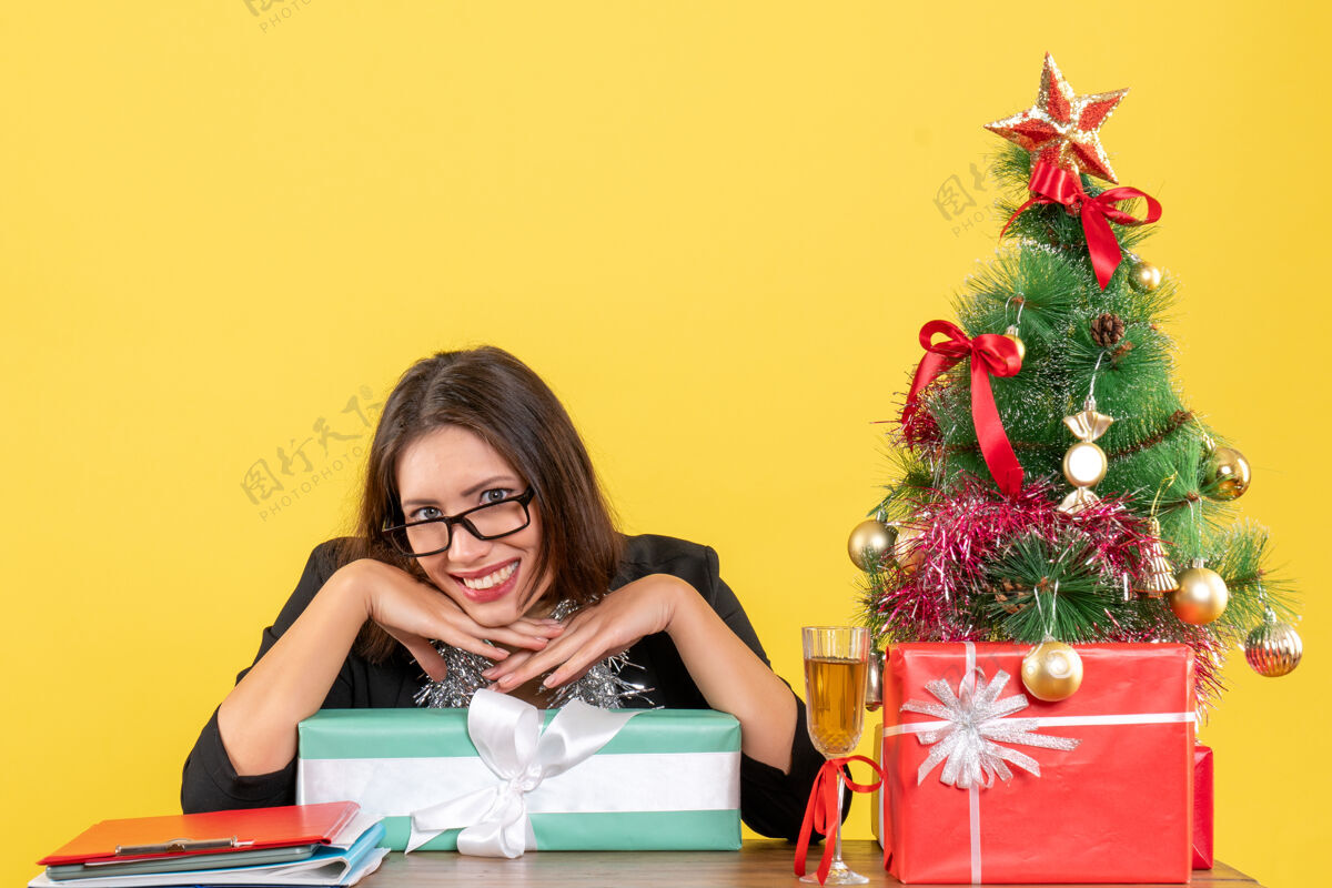 镜头微笑的商务女士穿着西装 戴着眼镜 展示着她的礼物 坐在一张桌子旁 办公室的镜头里有一棵圣诞树桌子漂亮眼镜