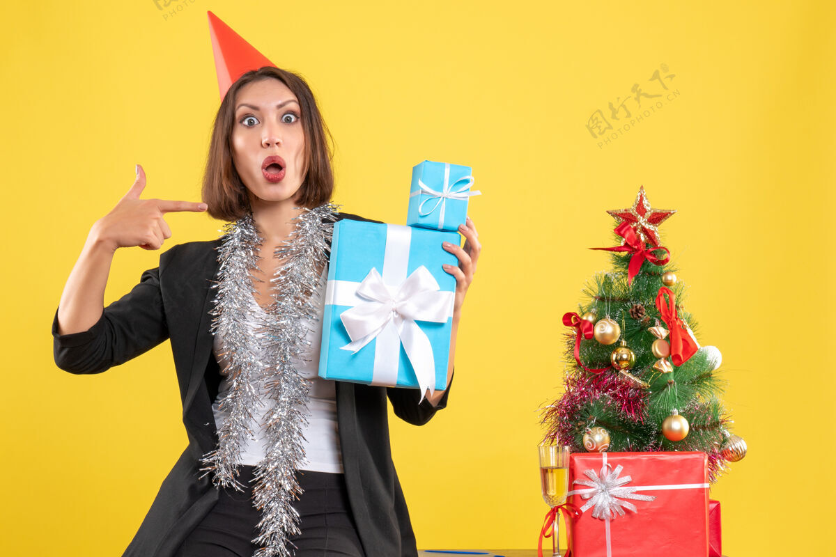 肖像圣诞节心情动情美丽的女士戴着圣诞帽指着黄色的办公室礼物礼物人漂亮