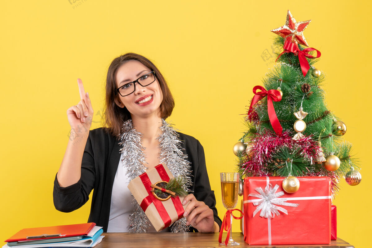 快乐在黄色的办公室里 一位穿着西装 戴着眼镜的漂亮商务女士坐在一张桌子旁 桌子上有一棵圣诞树眼镜花束西装