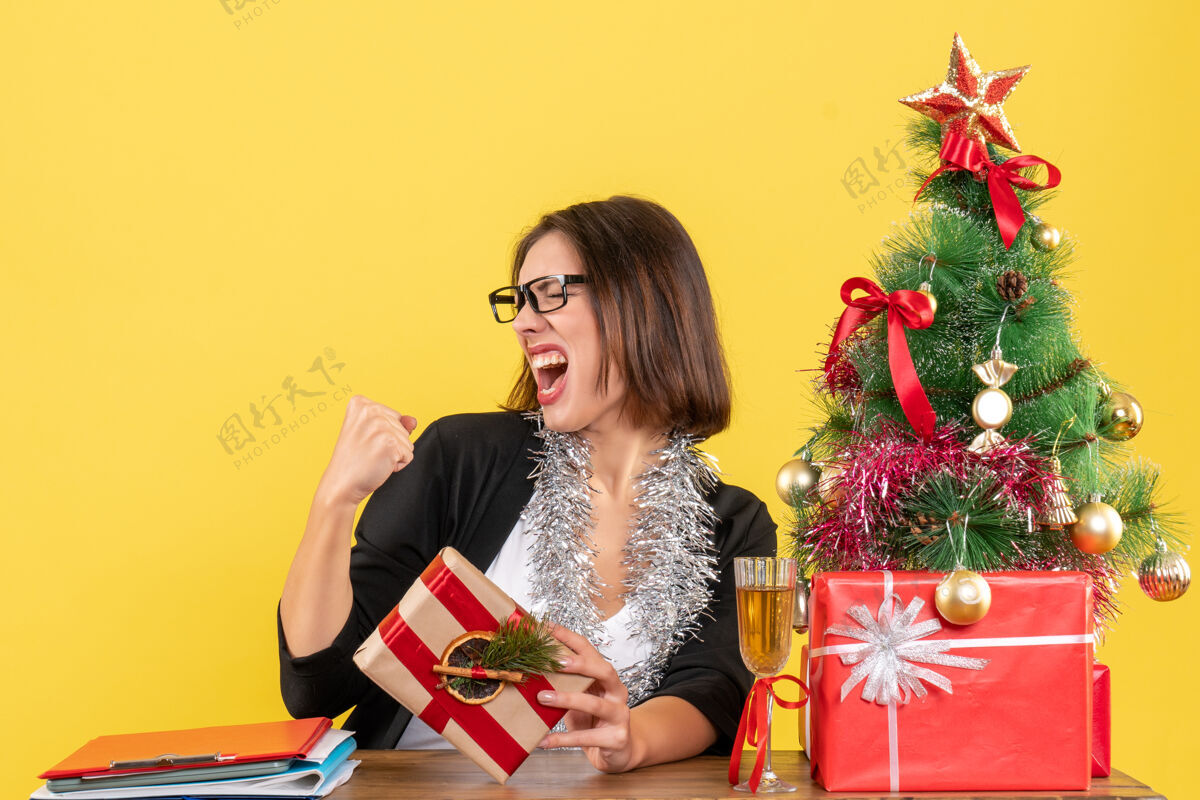 拿着一位穿着西装 戴着眼镜 骄傲地拿着礼物的美丽商务女士坐在一张桌子旁 桌子上放着一棵圣诞树快乐漂亮布置