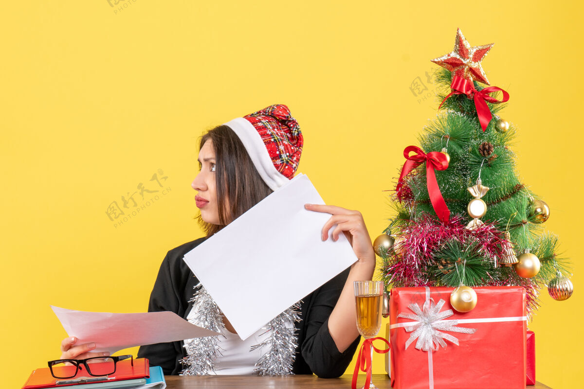 生活穿着西装 戴着圣诞老人帽和新年装饰品的商务女士感到困惑 坐在一张桌子旁 桌子上放着圣诞树困惑快乐感情