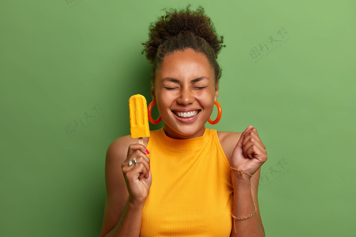 甜点快乐的黑皮肤千禧一代女孩吃着芒果味的黄色冰淇淋 紧握拳头期待着可怕的事情发生 有着牙齿般的微笑 穿着鲜艳的夏装 与世隔绝的绿色乐观可口卷发