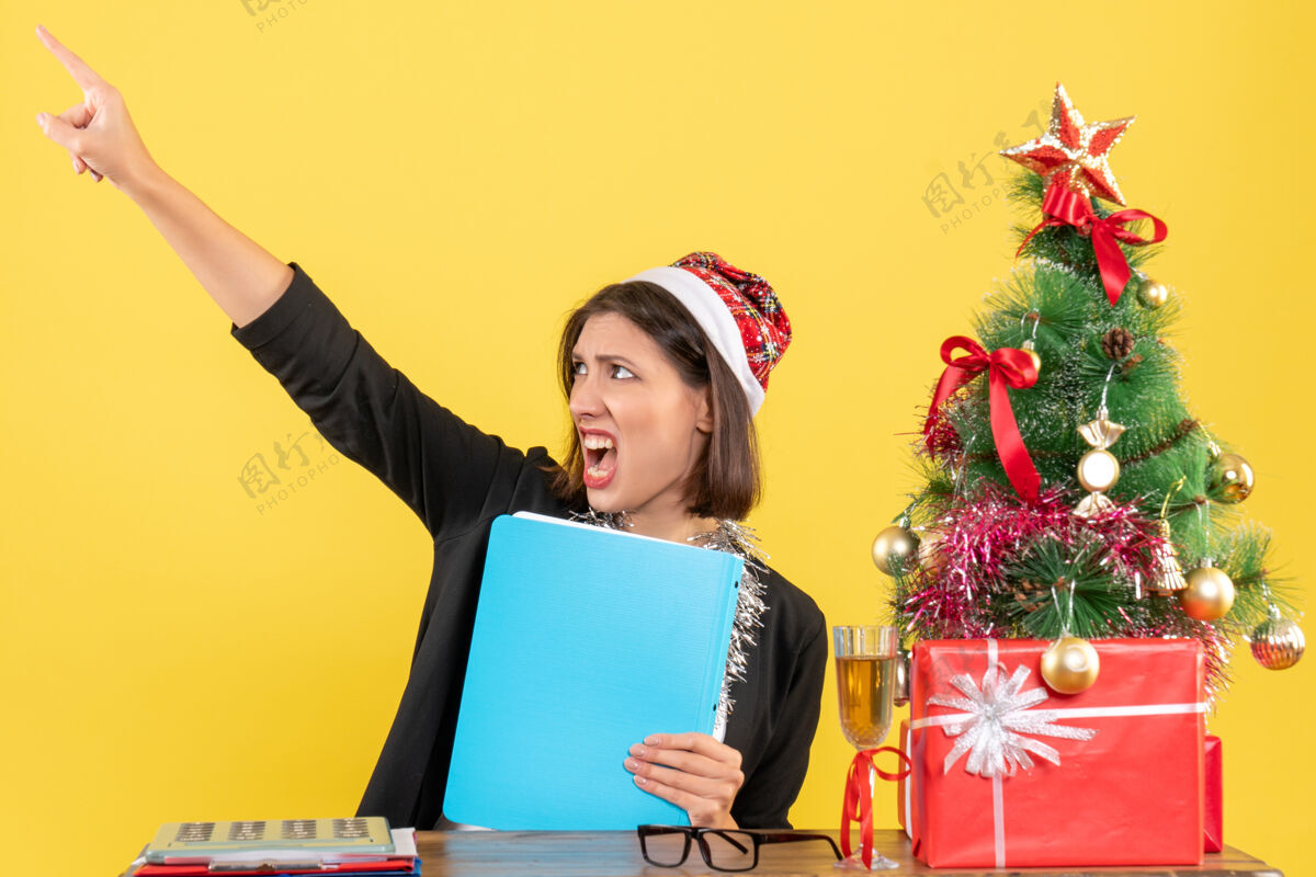 电脑迷人的女士穿着西装 戴着圣诞老人的帽子和新年装饰品 手里拿着文件指着办公室里的黄色隔离带指着黄色圣诞老人