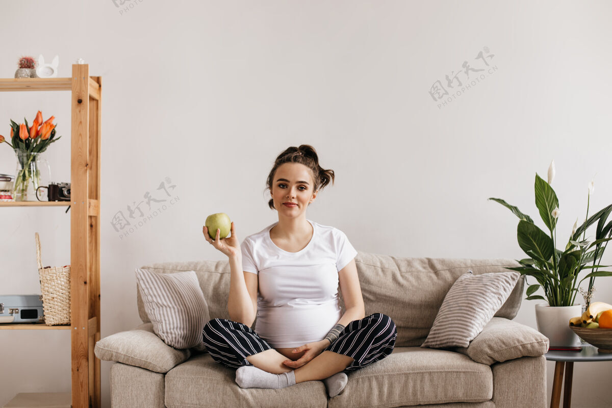 人穿着白色T恤和条纹裤的年轻孕妇手持青苹果腹部休息产妇
