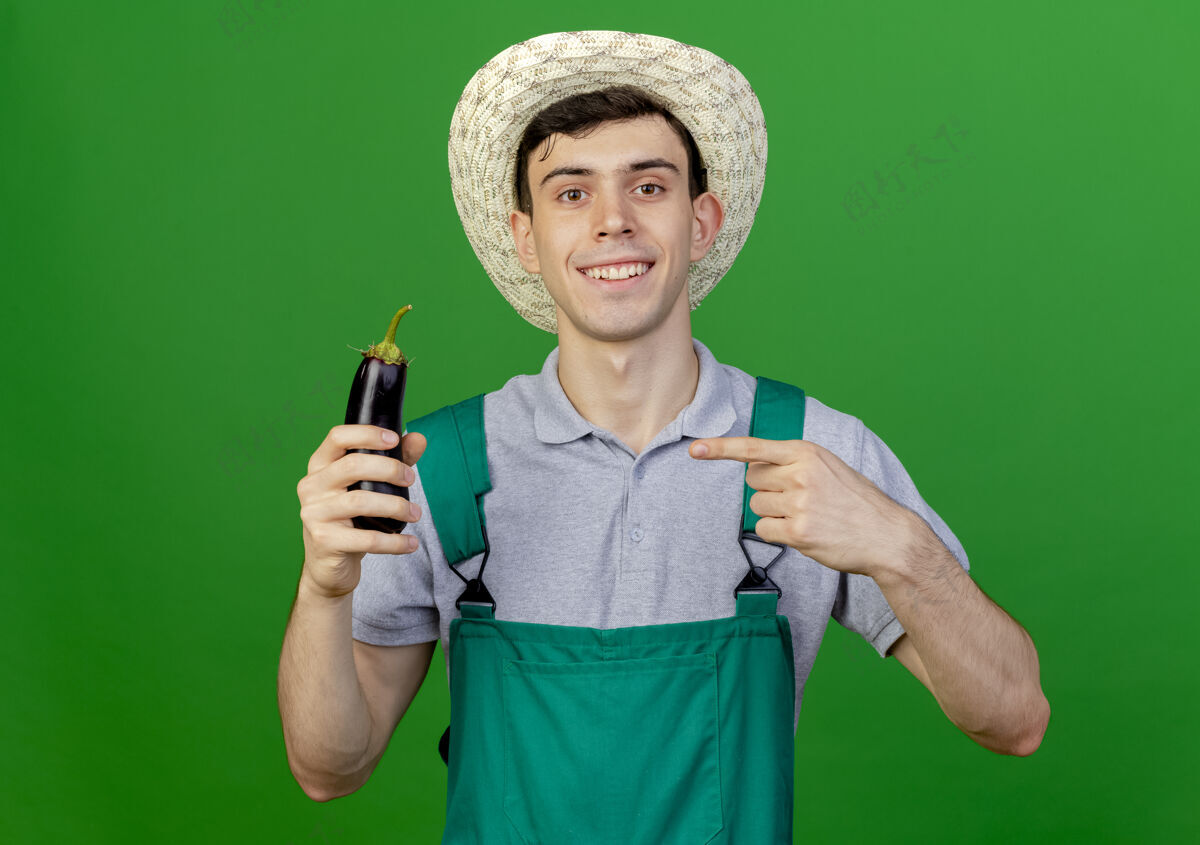 年轻戴着园艺帽的年轻男园丁微笑着 手拿茄子 指着隔离在绿色背景上的复制空间微笑园艺帽子