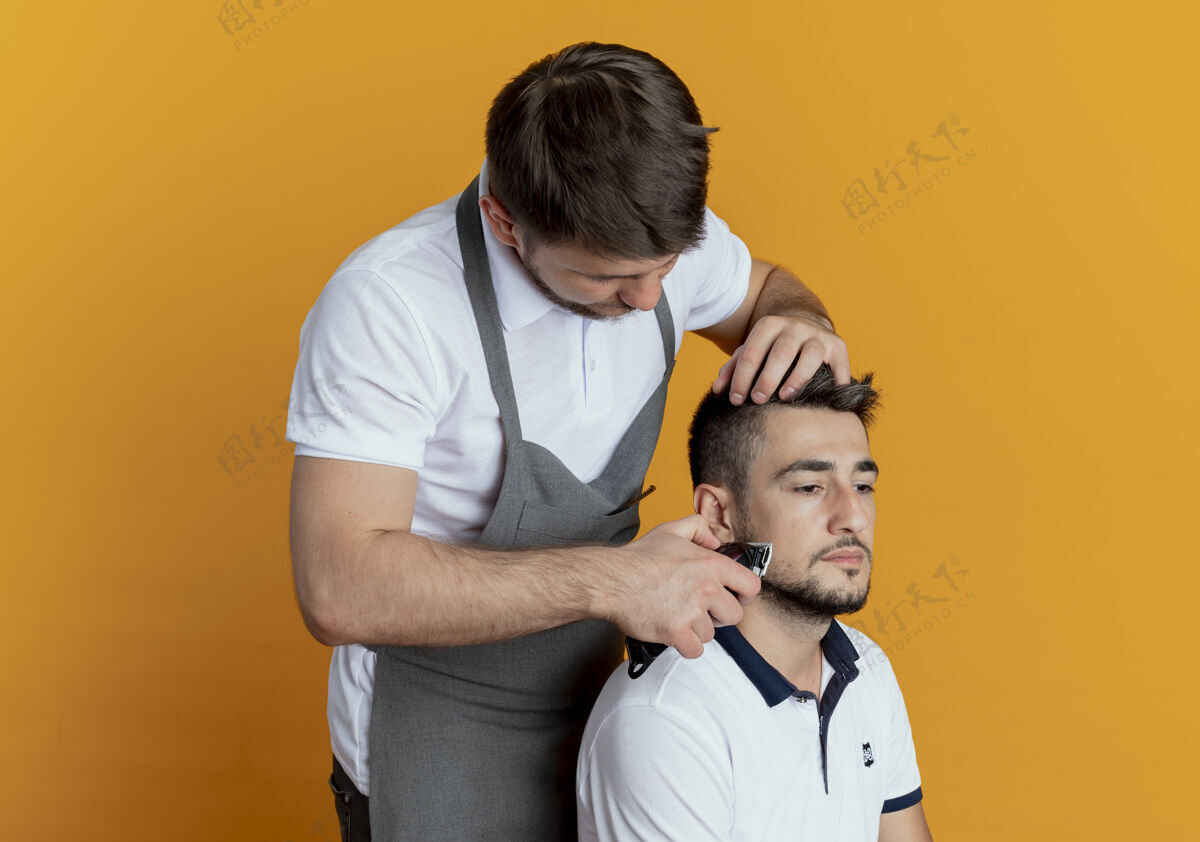 胡须穿着围裙的理发师用满意客户的剃须刀在橙色背景上刮胡子围裙满意修剪者