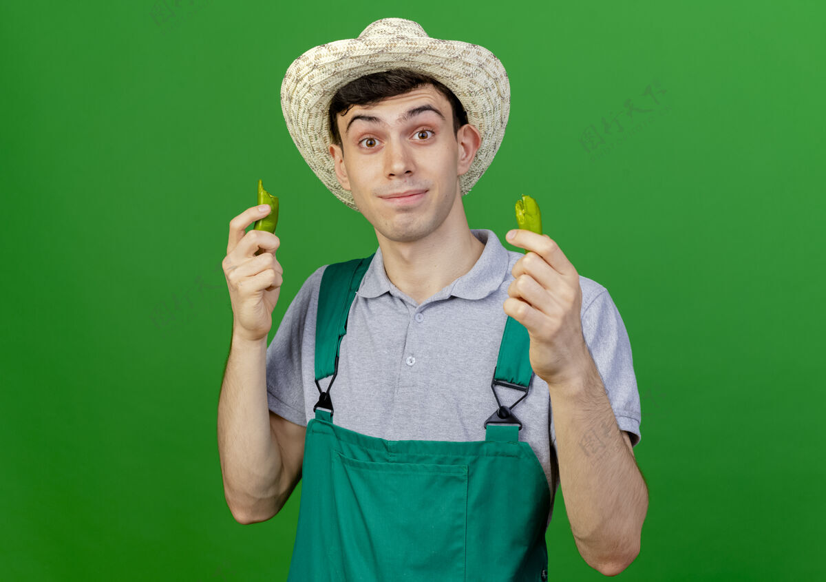 空间惊讶的年轻男园丁戴着园艺帽拿着破碎的辣椒看着相机隔离在绿色背景与复制空间帽子绿色花园