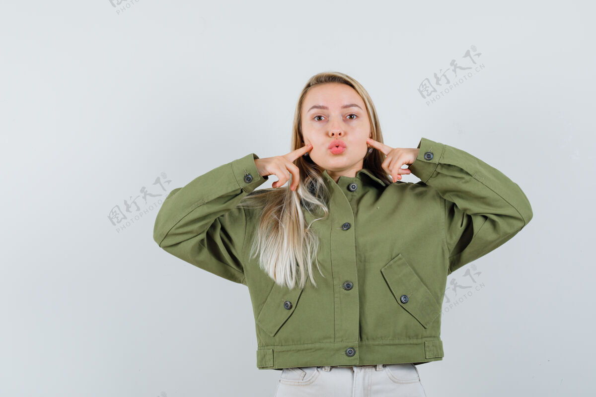 夹克穿着绿色夹克的年轻女性 牛仔裤指着她的脸颊 撅着嘴唇 正面视图脸头发嘴唇