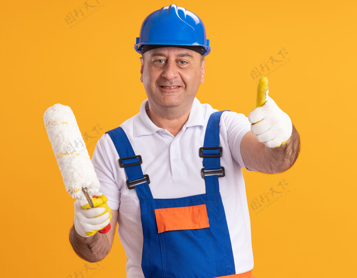 脸身着制服 戴着防护手套 面带微笑的成年建筑工人竖起大拇指 拿着隔离在橙色墙上的滚筒刷人穿着成人