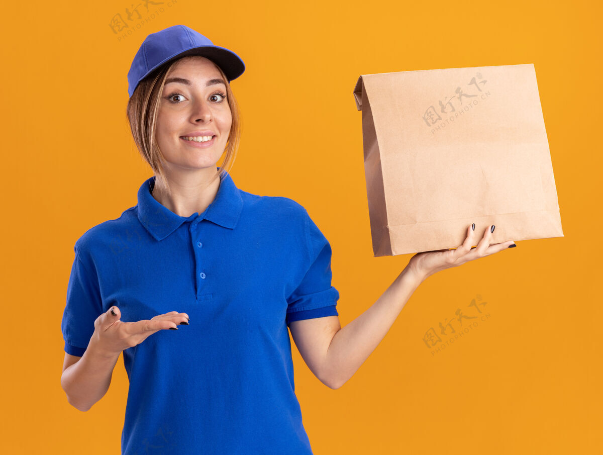 人令人印象深刻的年轻漂亮的女送货员在制服举行和点在纸包装与手隔离在橙色墙上衣服感情包装