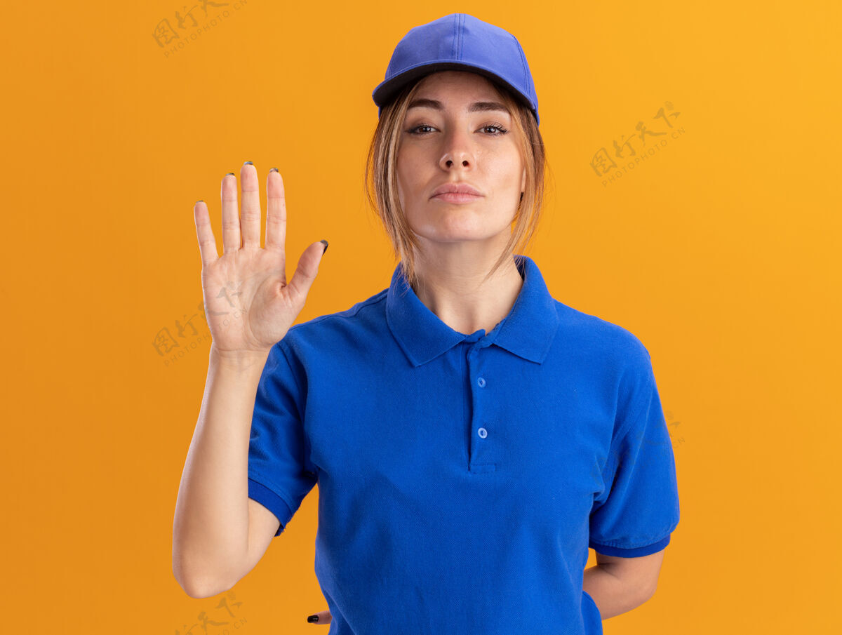 姿势自信的年轻漂亮的女送货员穿着制服站在橙色的墙上 举手孤立手脸人
