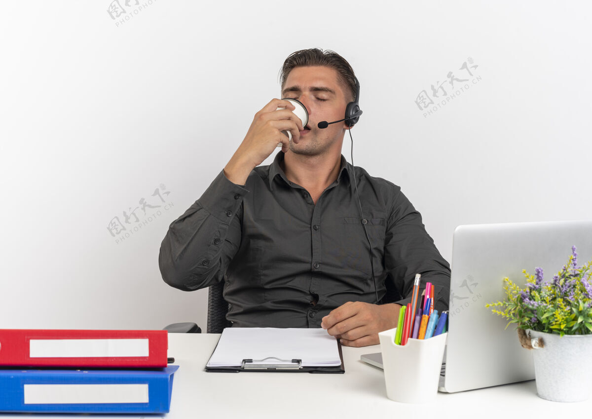 年轻年轻而快乐的金发上班族戴着耳机坐在办公桌旁 拿着办公工具 用笔记本电脑 喝着一杯咖啡 背景是白色 有复印空间使用金发咖啡