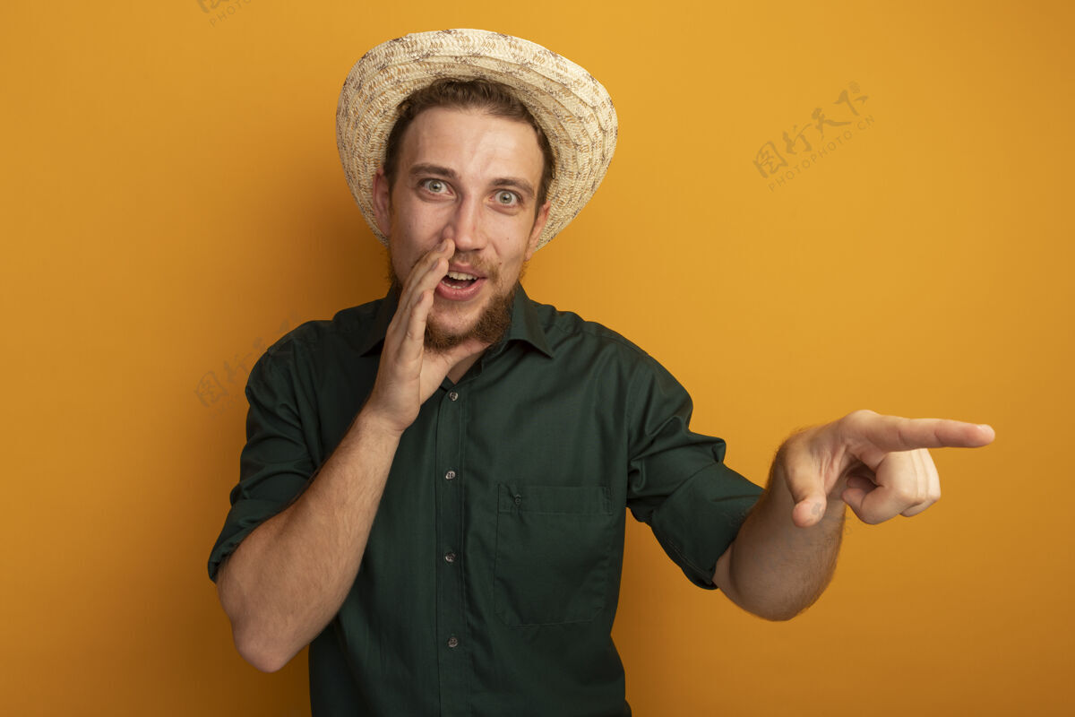 欢乐快乐的金发帅哥 戴着沙滩帽 手紧挨着嘴 指着橘色墙上孤立的一面人站衣服