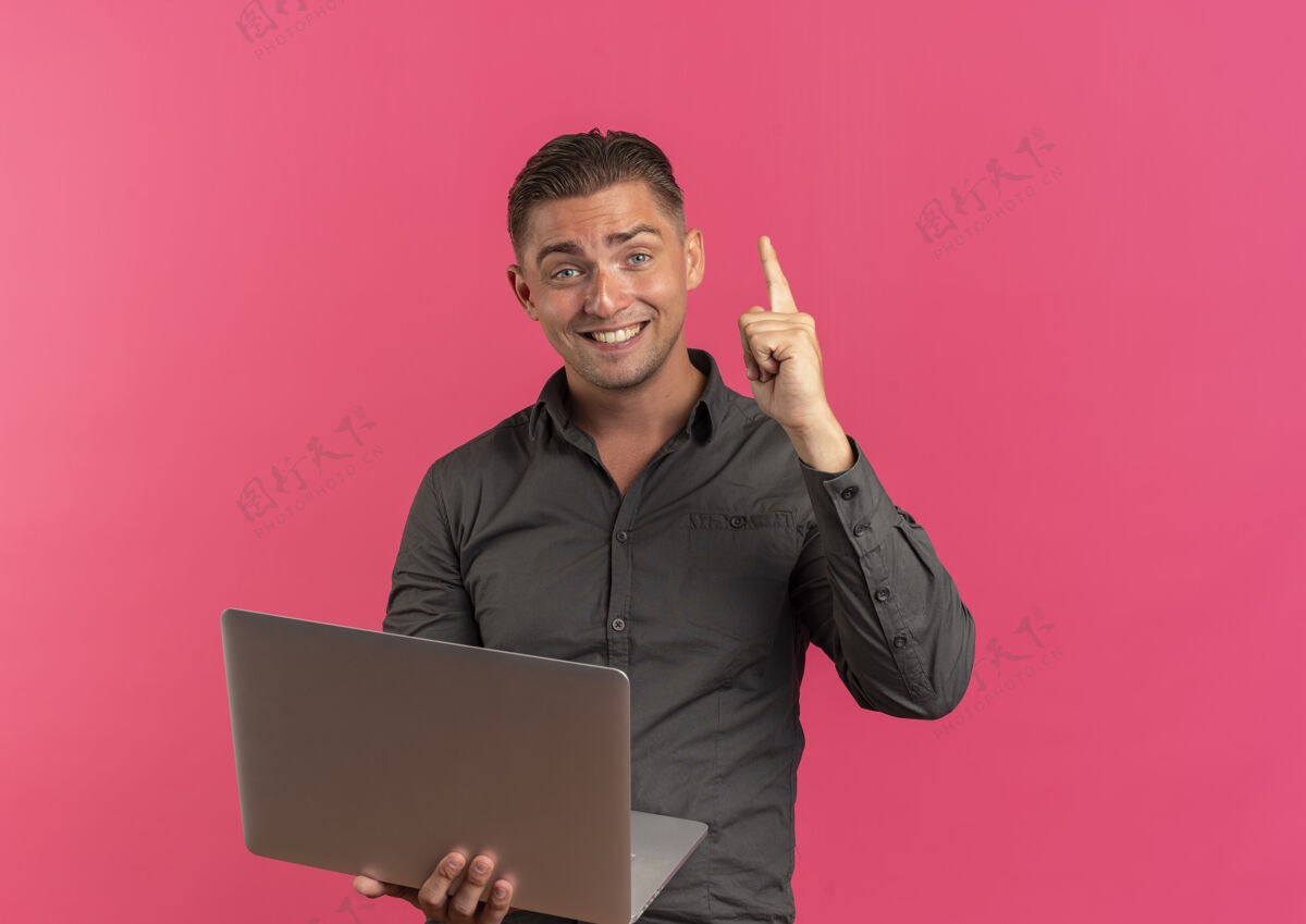笔记本电脑年轻的金发碧眼帅气的男子手持笔记本电脑 抬头看着隔离在粉色背景上的相机 还有复印空间年轻相机英俊