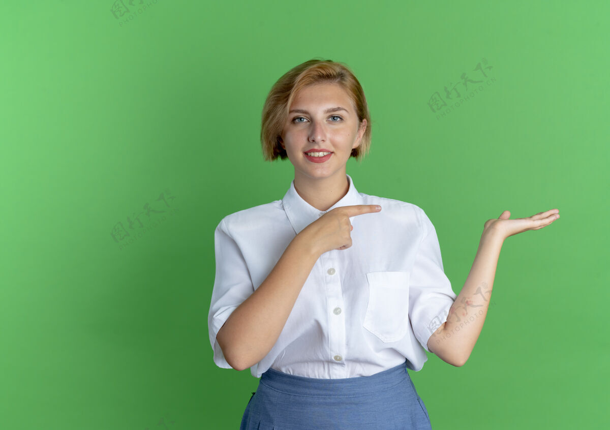 年轻年轻的微笑着的金发俄罗斯女孩指着被隔离在绿色背景上的空白的手手女孩金发