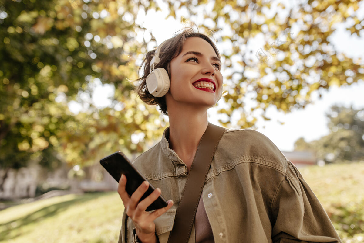 成人乐观的女人 深色头发 穿着橄榄色牛仔服 微笑着拿着电话戴着轻型耳机的女人在户外摆姿势音乐城市手机