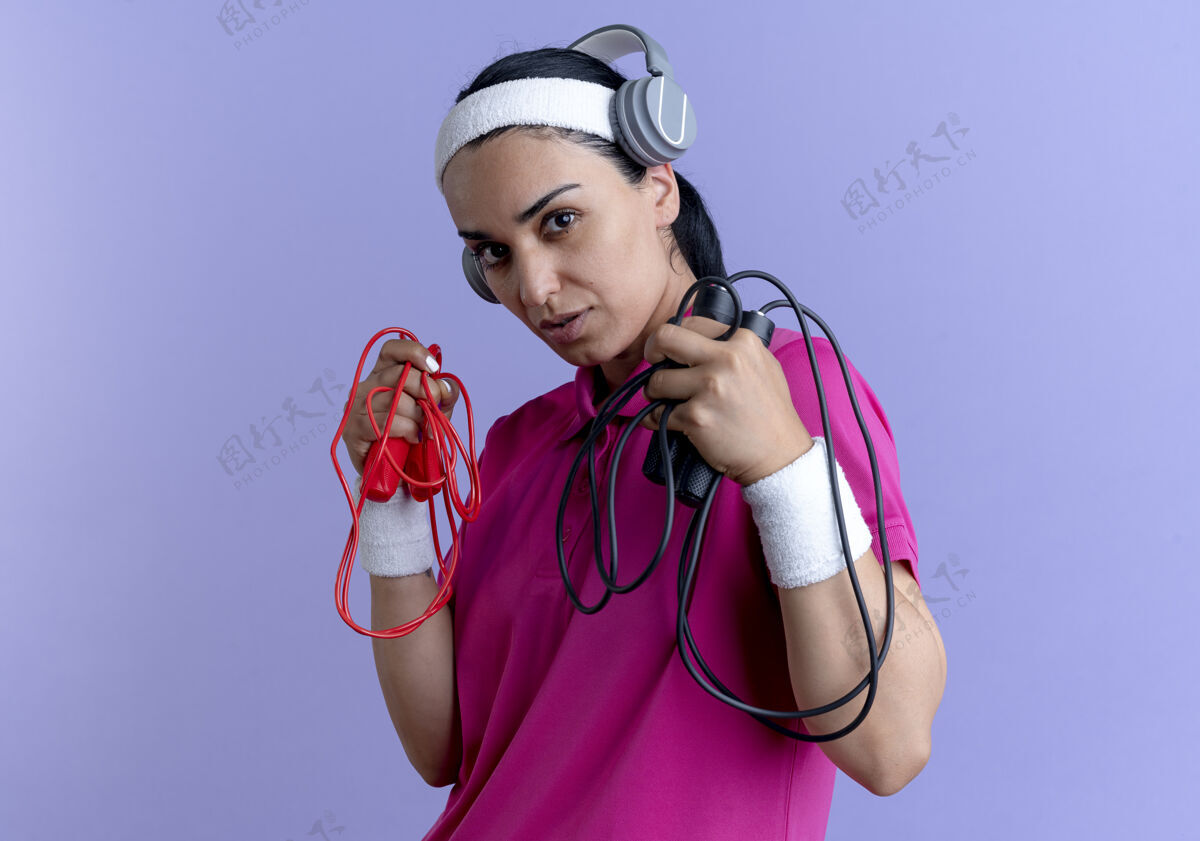 耳机年轻自信的白人运动女性戴着头带 戴着耳机戴着腕带 手里拿着跳绳 紫色背景上有复制空间头带佩戴腕带