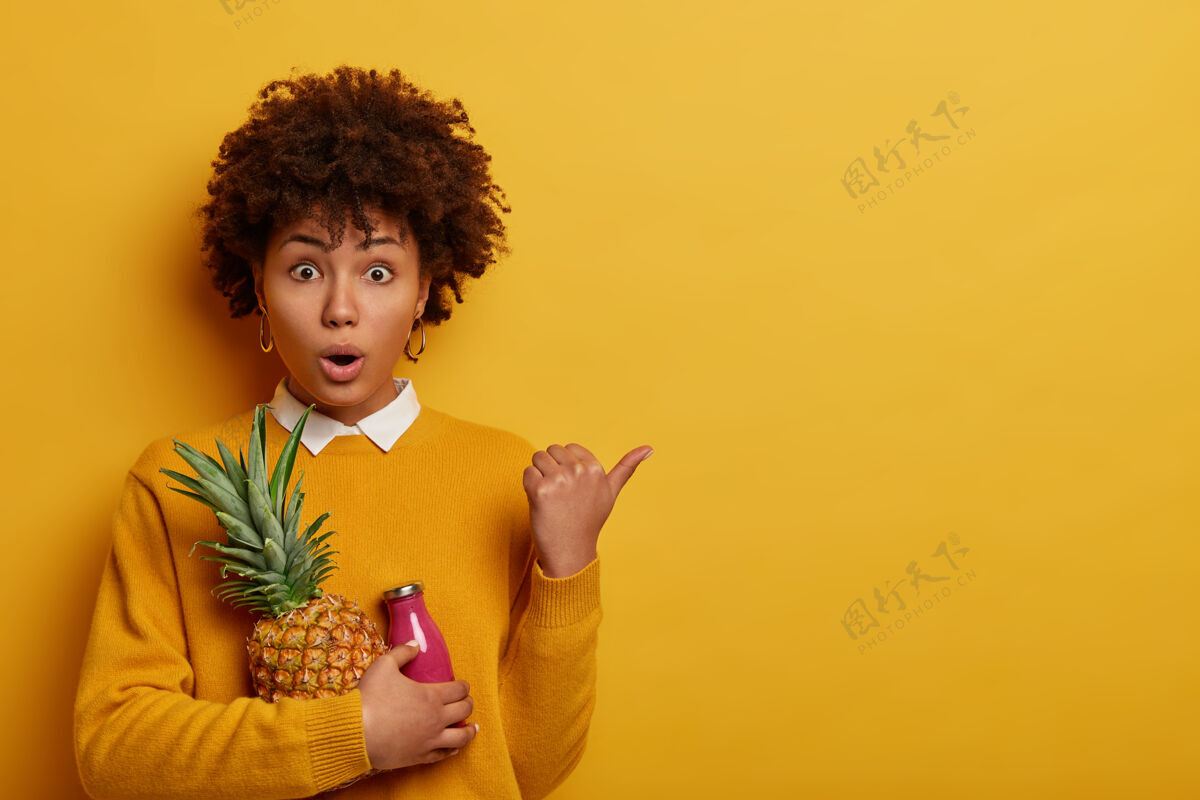 卷曲一个非洲头发的女人拿着新鲜的菠萝和一瓶粉红色的奶昔 很惊讶 很有感情奇迹旁白拇指