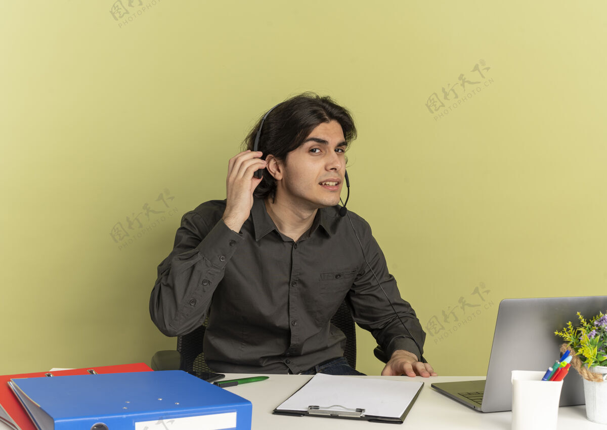 工人戴着耳机的年轻上班族坐在办公桌旁 拿着笔记本电脑的办公工具 拿着耳机 试图在绿色背景和复印空间中听到孤立的声音烦恼坐着尝试