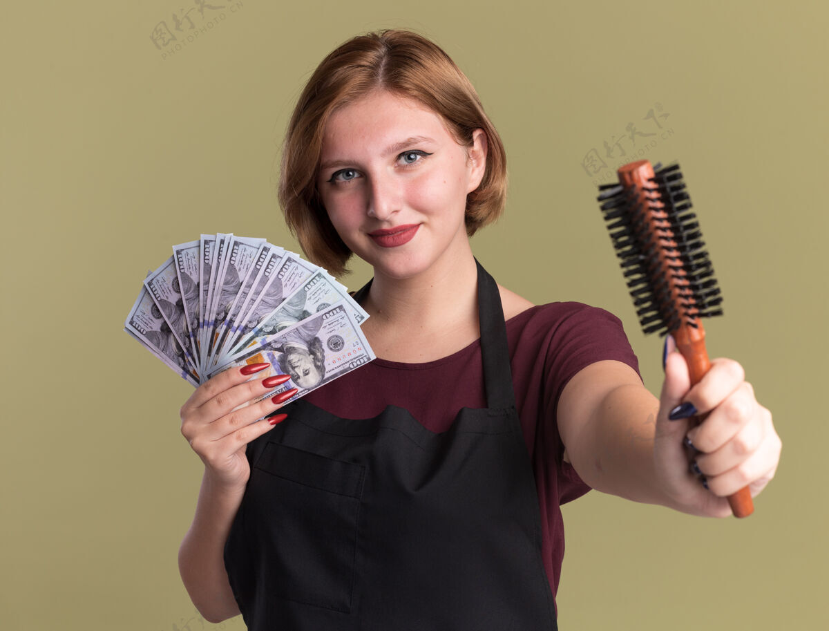 展示年轻漂亮的女理发师围着围裙拿着现金展示着发刷微笑自信地站在绿色的墙上头发理发师刷子