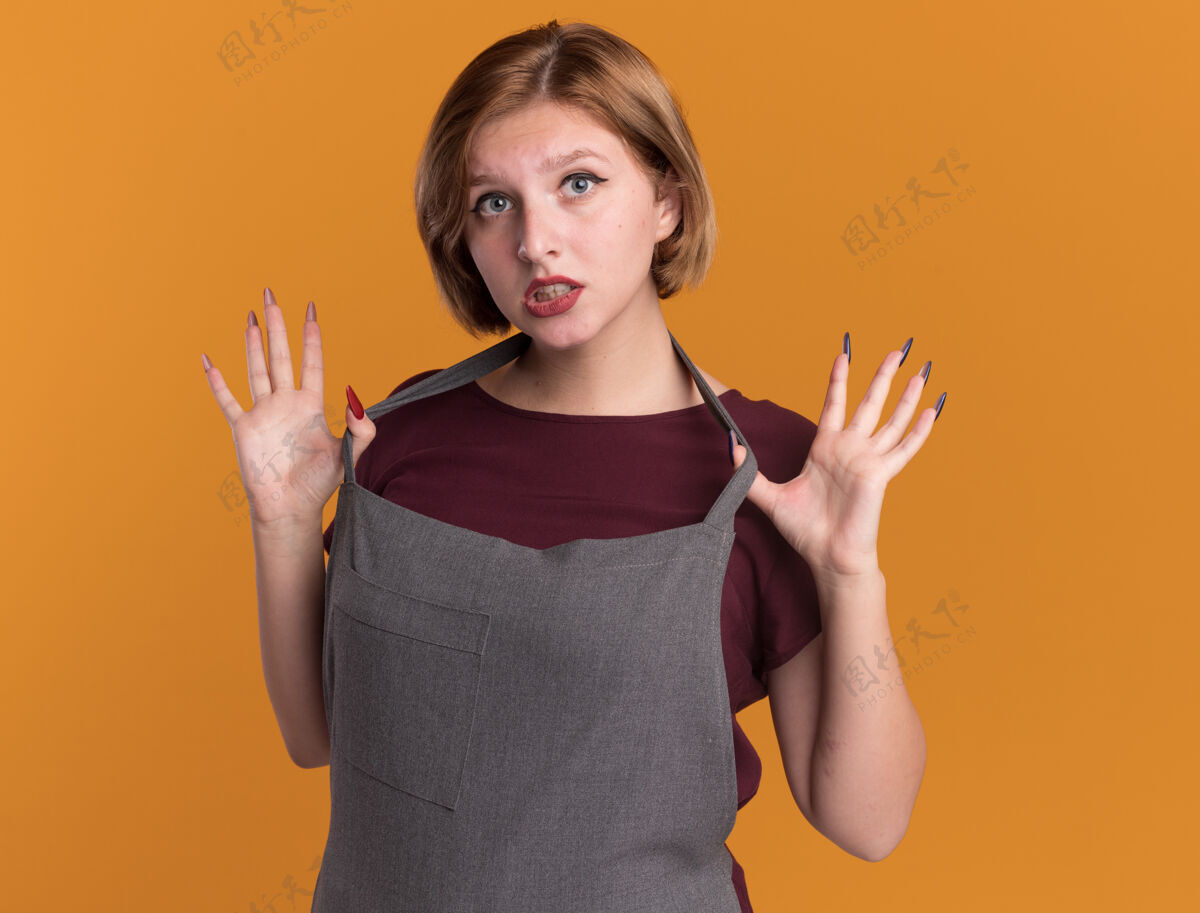 漂亮年轻漂亮的女理发师穿着围裙望着前面迷茫的手臂抬起疑惑的站在橘色的墙上围裙手臂怀疑