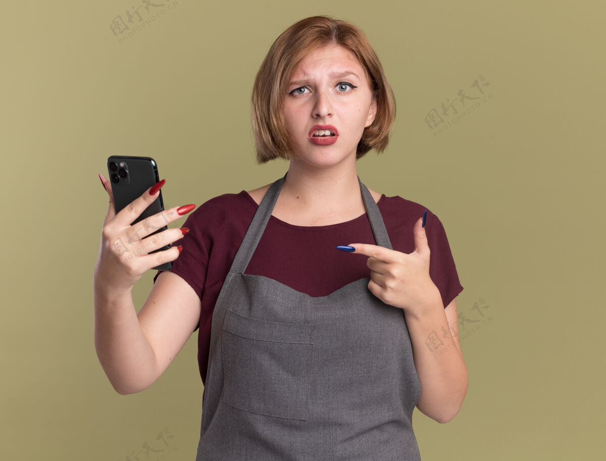 年轻年轻漂亮的女理发师围着围裙 拿着智能手机 用食指指着站在绿色墙壁上困惑的它站着移动困惑