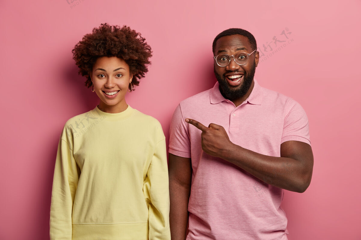 Indexfinger穿粉色t恤的快乐男人的水平镜头指向女友 拥有积极的表情套头衫高兴男人