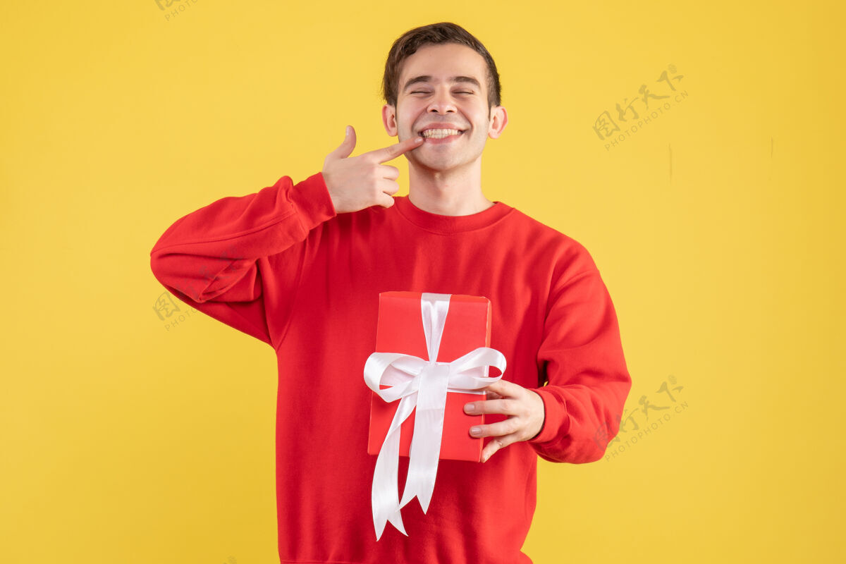 视图正面图：年轻人拿着红色礼盒指着黄色背景上的微笑快乐人物衬衫