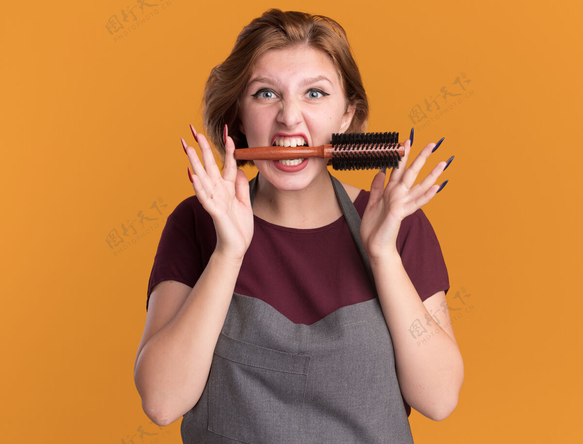 情绪年轻漂亮的女理发师围着围裙 嘴里叼着发刷 站在橘色的墙上 情绪激动地看着前面头发嘴巴女人