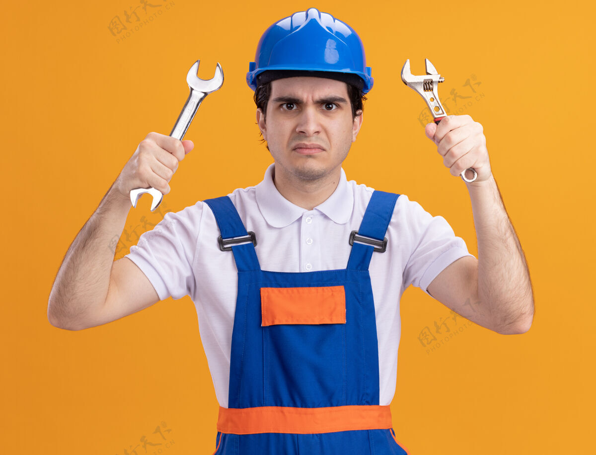 年轻年轻的建筑工人穿着建筑制服 戴着安全帽 拿着扳手 站在橙色的墙上怒气冲冲地看着前方脸愤怒男人