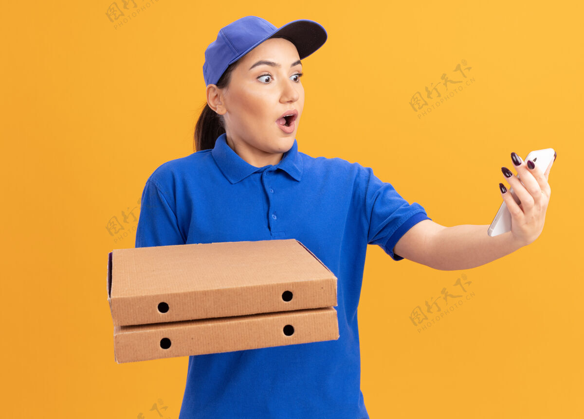 惊喜身穿蓝色制服 戴着帽子 拿着披萨盒的年轻送货员站在橙色的墙上 看着自己的智能手机 既惊讶又惊讶站着太棒了披萨