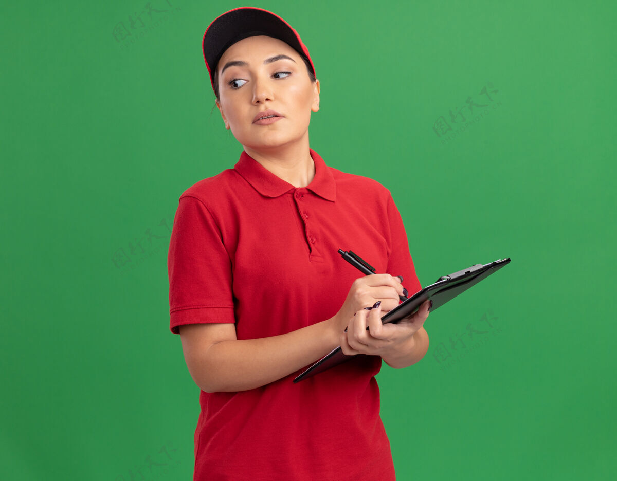 严肃身穿红色制服 头戴帽子的年轻女送货员拿着剪贴板和铅笔 带着严肃自信的表情站在绿色的墙边看着站着铅笔表情