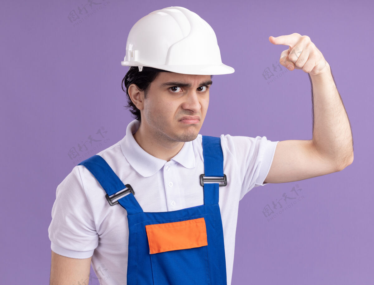 安全年轻的建筑工人穿着建筑制服 戴着安全帽 怒气冲冲地看着前面 食指指着前面 站在紫色的墙上脸愤怒年轻