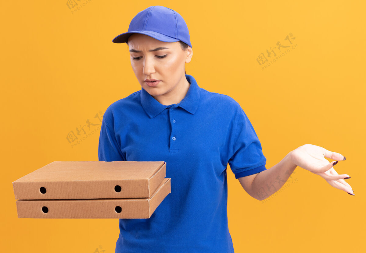 女人身穿蓝色制服 头戴鸭舌帽 拿着比萨饼盒的年轻女送货员站在橙色的墙上 看着他们困惑不解困惑年轻拿着