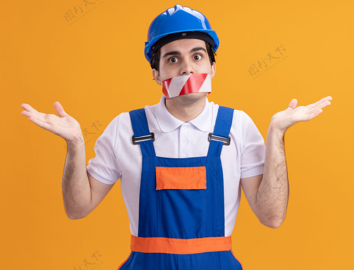 头盔年轻的建筑工人穿着建筑制服 戴着安全帽 嘴上缠着胶带 困惑地耸耸肩站在橙色的墙上站着建设者围着