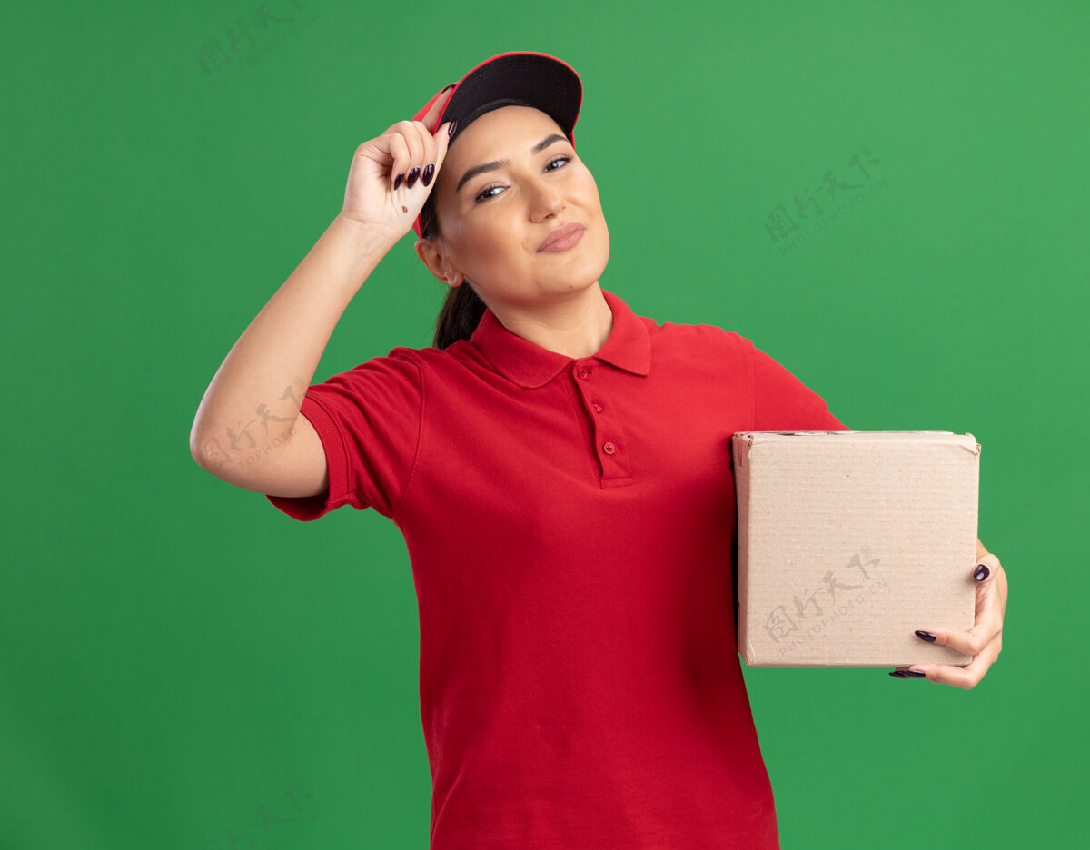 立场年轻的送货员身穿红色制服 戴着帽子 拿着纸板箱 站在绿色的墙上 自信地微笑着看着前面帽子盒子信心