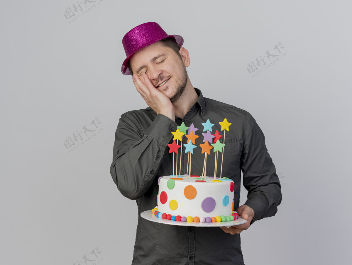 拿着高兴的年轻人闭着眼睛戴着粉红色的帽子拿着蛋糕把手放在脸上孤立的白色背景眼睛手帽子
