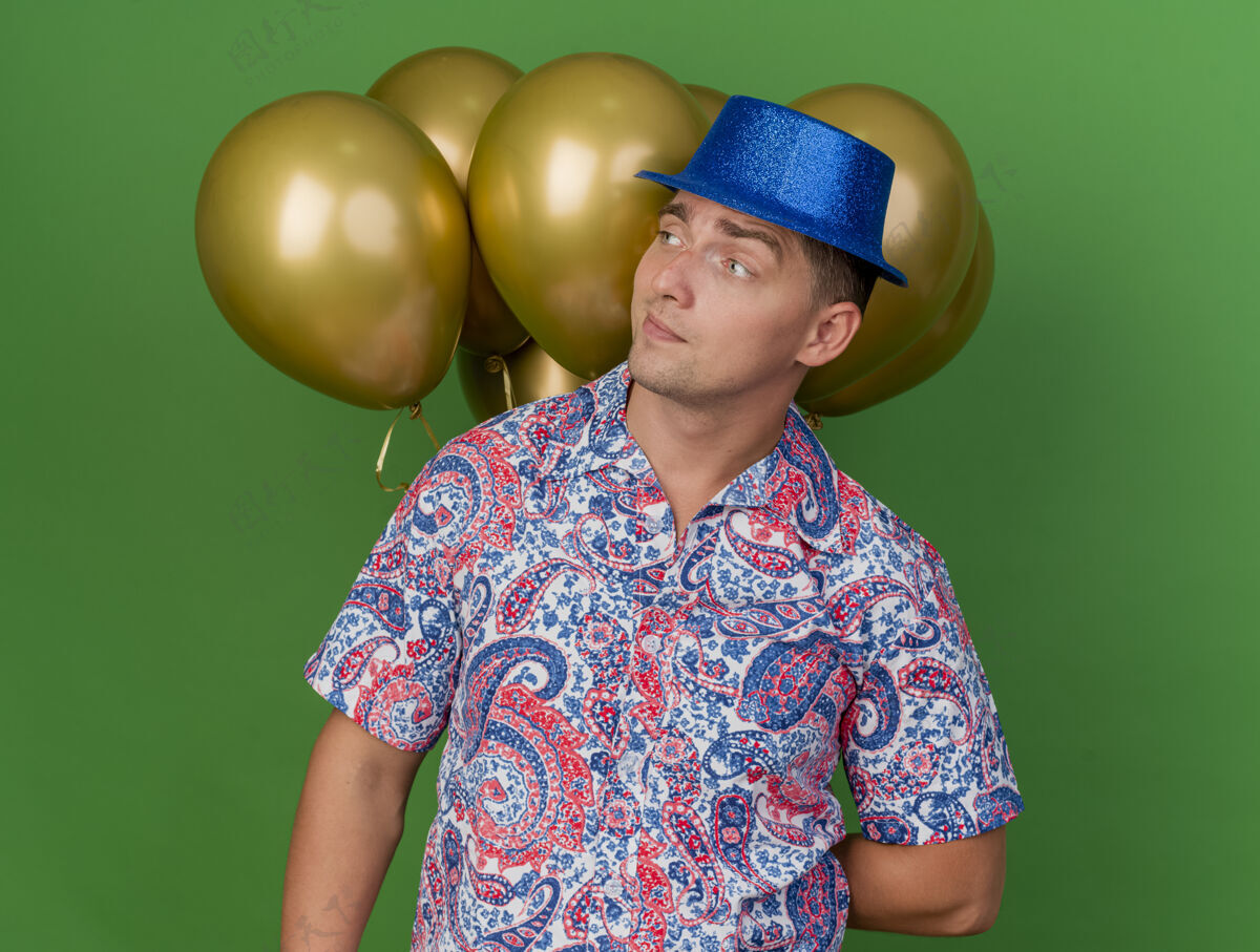 年轻人令人印象深刻的年轻人看在一边戴着蓝色帽子站在气球前孤立的绿色背景气球站帽子