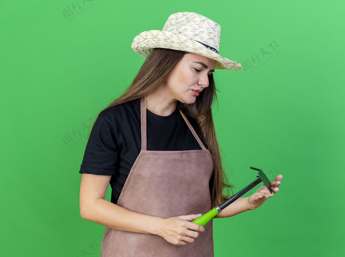 锄头想着美丽的园丁 穿着制服的女孩戴着园艺帽 拿着锄头 看着绿色背景上孤立的耙子花园举行制服