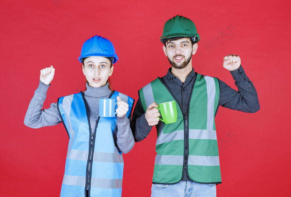 成功男女工程师戴头盔 手持蓝绿色马克杯 并显示满意标志年轻提升人类
