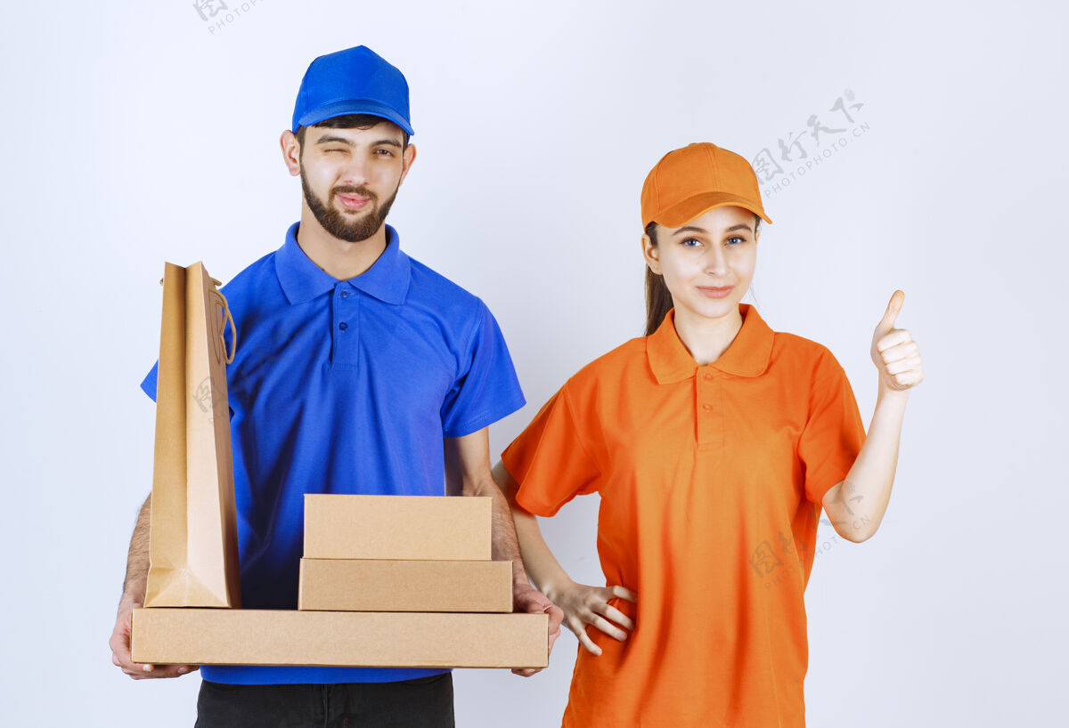 美食家身穿蓝黄制服的男女快递员手持纸板外卖盒和购物包 并展示满意手势员工工人享受