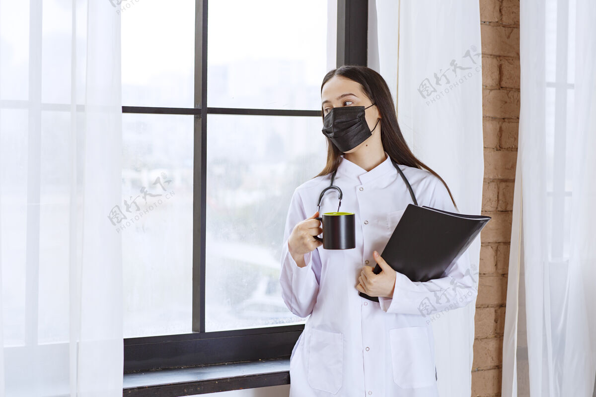 细菌医生拿着听诊器和黑色面罩 手里拿着一杯黑色饮料和一个黑色文件夹 从窗户往外看咖啡消毒诊所