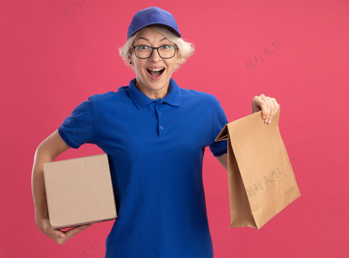 女人身穿蓝色制服 头戴鸭舌帽的年轻女送货员手里拿着纸包和纸箱 在粉红色的墙上高兴而兴奋年轻抱着包装